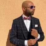 Image of Yahya Pandor, Gloucestershire SLC member. He is standing against an orange wall, looking to the side. he is wearing sunglasses, a suit and a red bow tie.