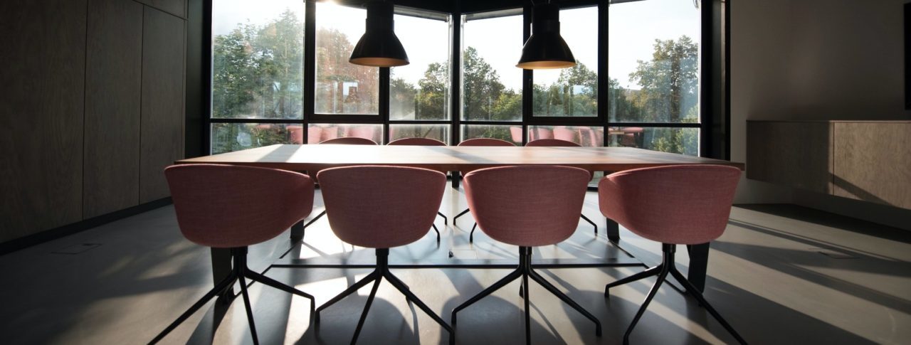 Image shows a rectangular table with four orange chairs tucked underneath. There are two large lights hanging over the table. The table is in front of a large window.