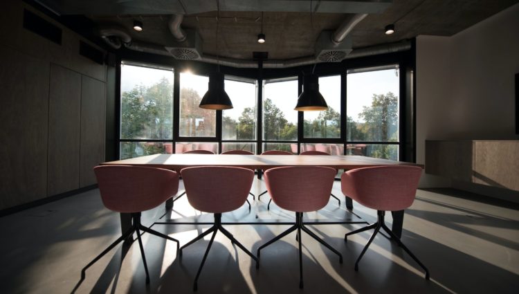 Image shows a rectangular table with four orange chairs tucked underneath. There are two large lights hanging over the table. The table is in front of a large window.