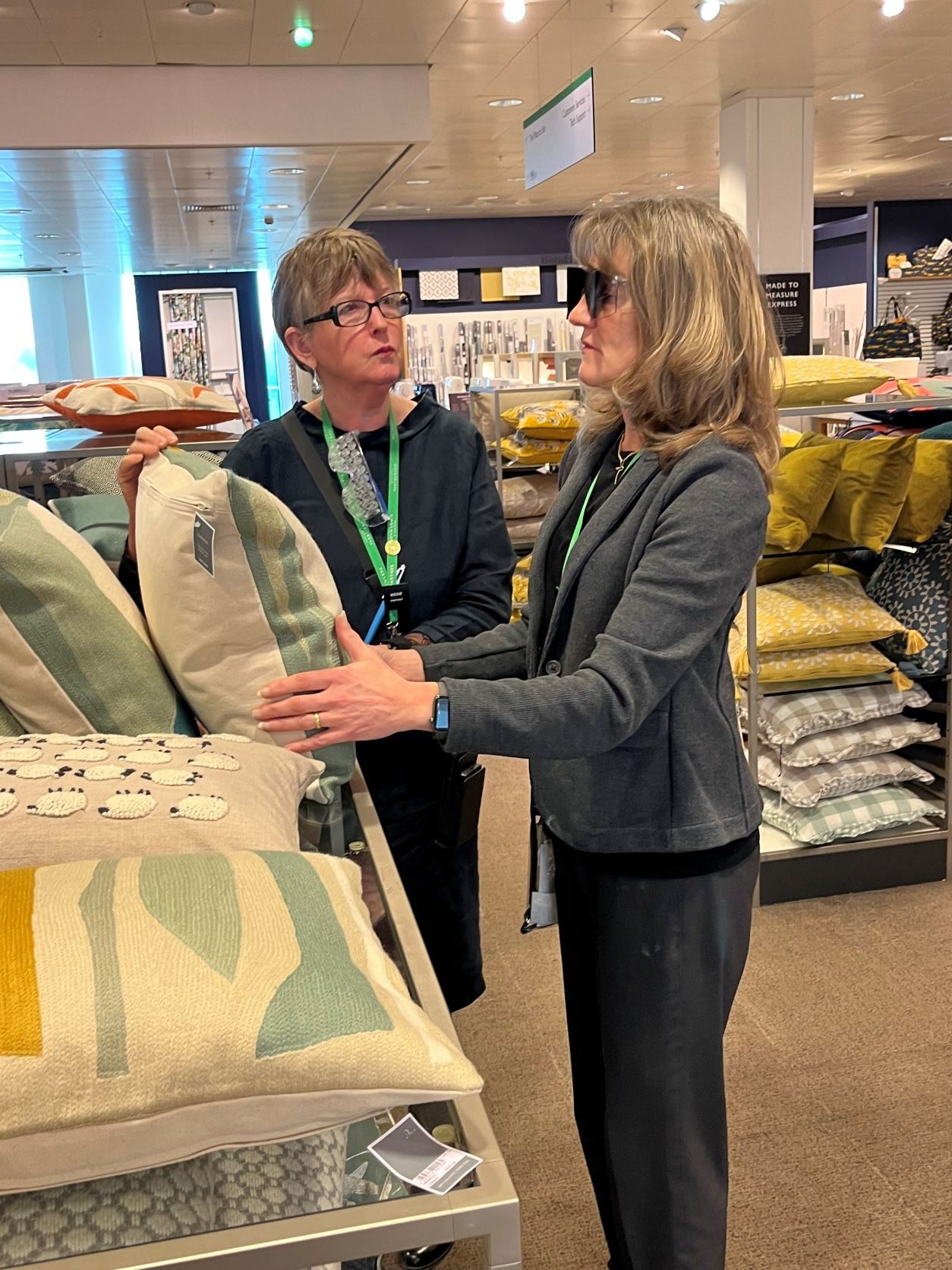 Two John Lewis staff members seen on the shop floor, looking at cushions. One staff member is wearing sim specs, the other is guiding.
