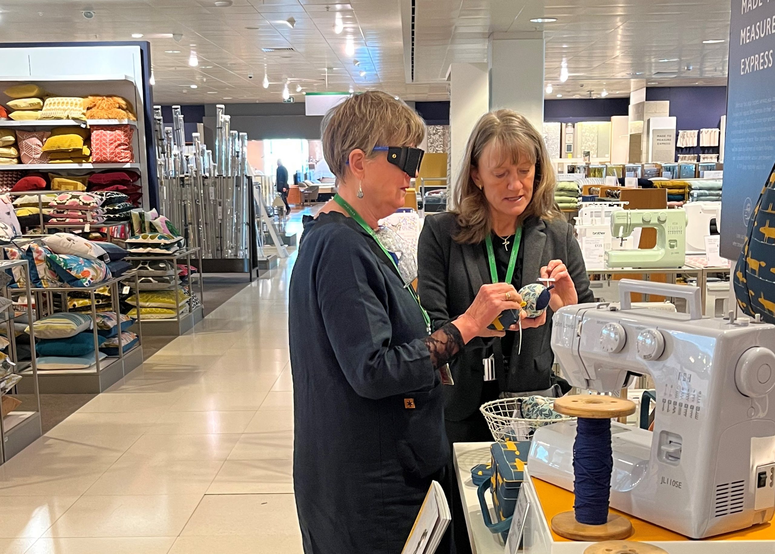 Two John Lewis staff members looking at items in the kitchen department. One is wearing sim specs and holding an item in her hand. The other is guiding and assisting her.
