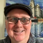 Head shot of Pete Hoey, North Yorkshire SLC member. Pete is standing in front of a painting of an old building. He is wearing a flat cap and glasses, and smiling at the camera.