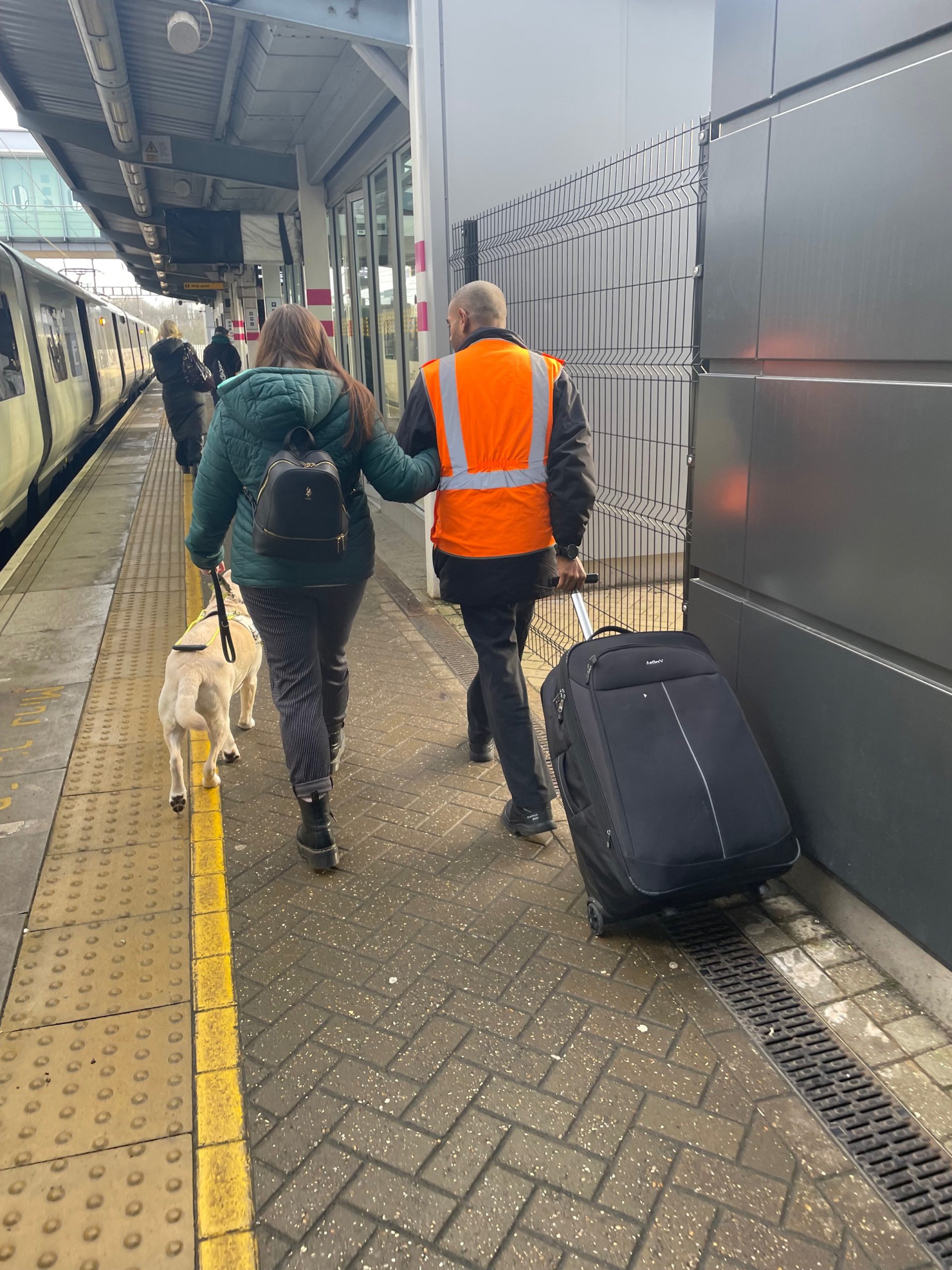 Image shows Samantha Leftwich, Bedfordshire Engagement Manager, being assisted by GTR staff at Luton Parkway. Sam is being guided by the arm and the assistant is pulling a large suitcase. Sam is holding guide dog lizzie’s lead. They are walking along the platform, next to a train. Their backs are to the camera.