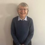 Yorl SLC member, Susan Dinkinson. Susan is leaning against a white wall, facing the camera, smiling. She is wearing glasses and has short, bobbed, hair.