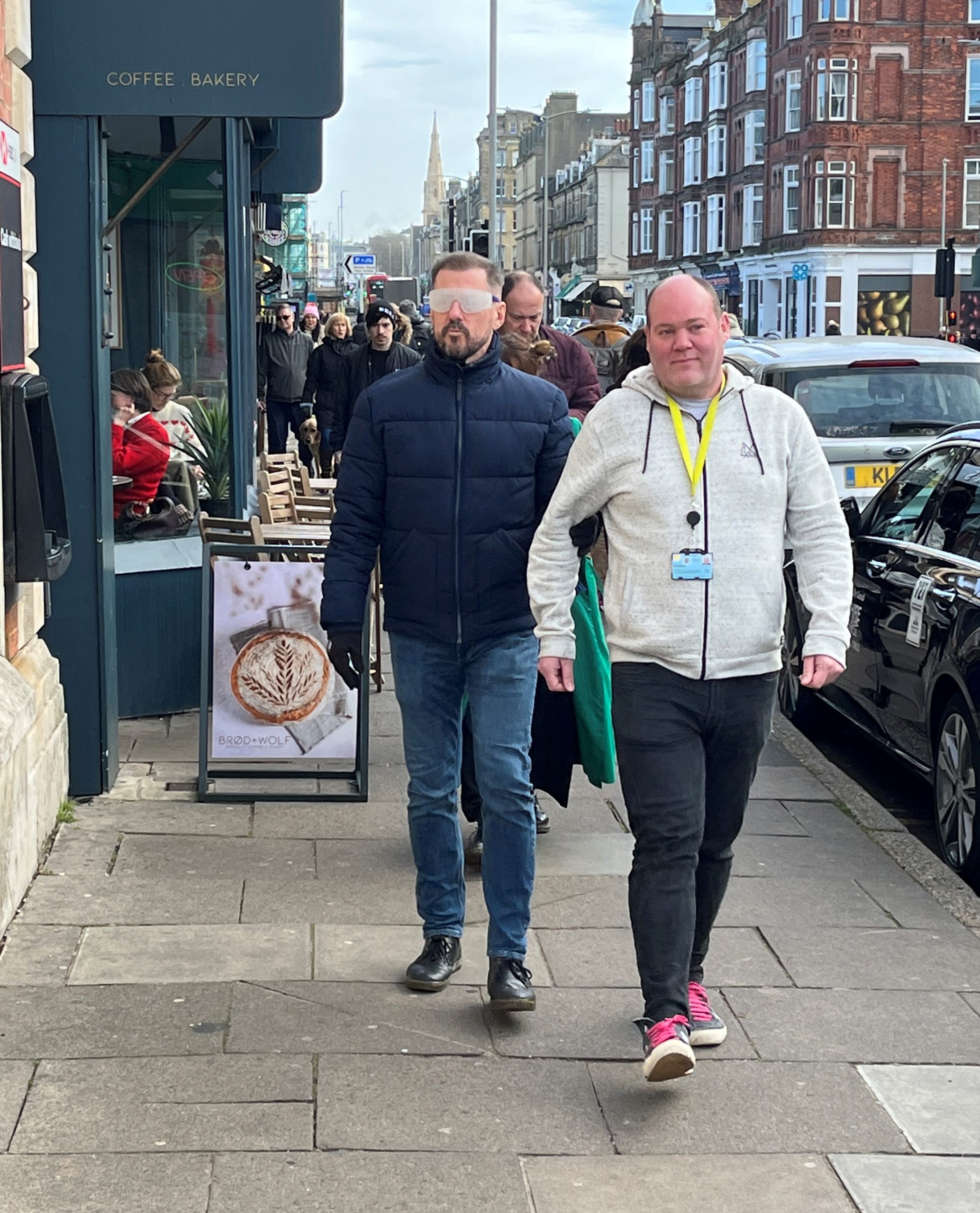 Two attendees from Brighton & Hove City Council on the sim spec walk. They are walking down a street with outdoor furniture seen outside a café. One man is wearing sim specs and being guided.