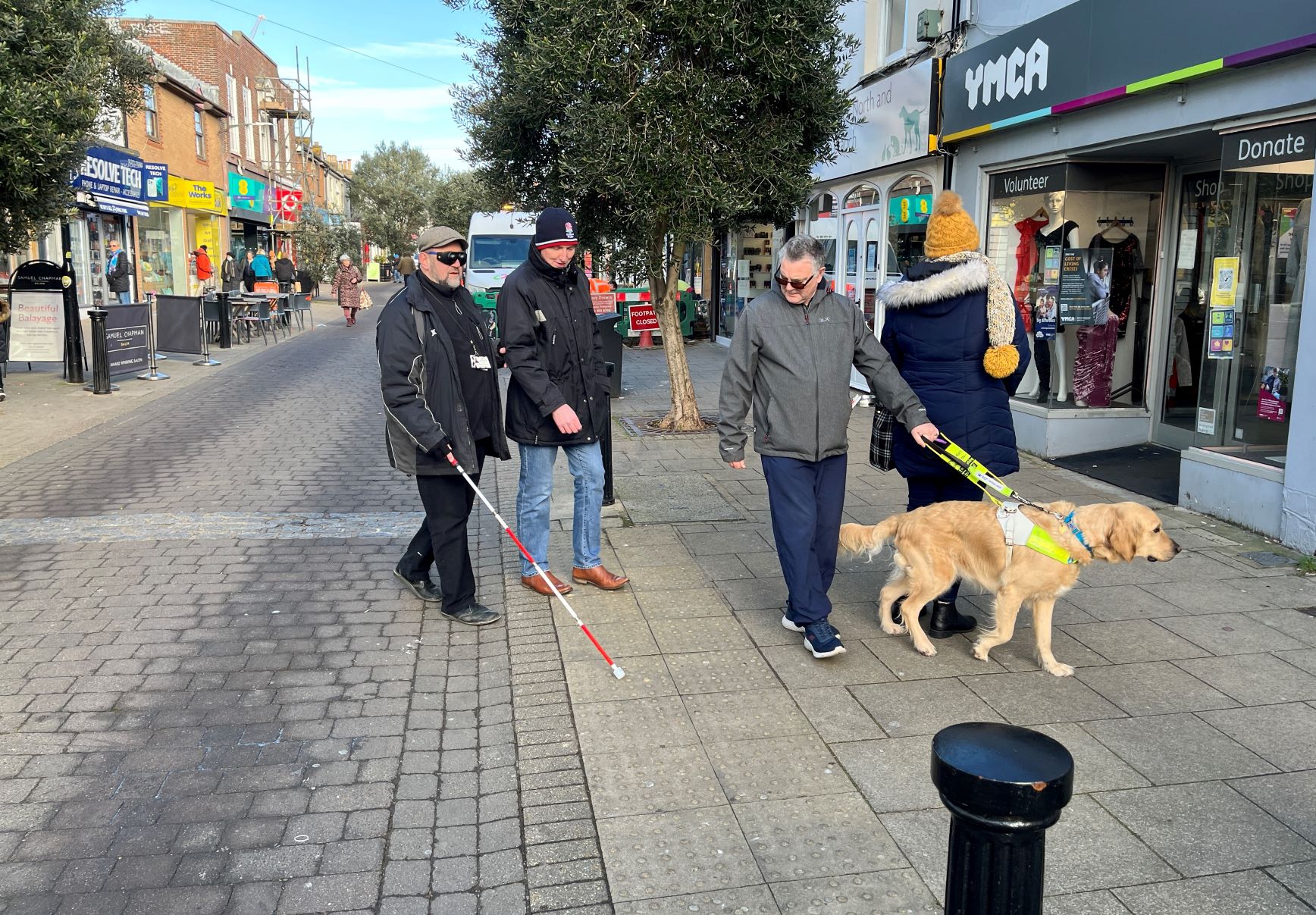 SLC member Graham and guide dog teddy, are with two attendees. One is wearing sim specs and walking with a cane, the other is guiding him. They are crossing a street lined with shops.