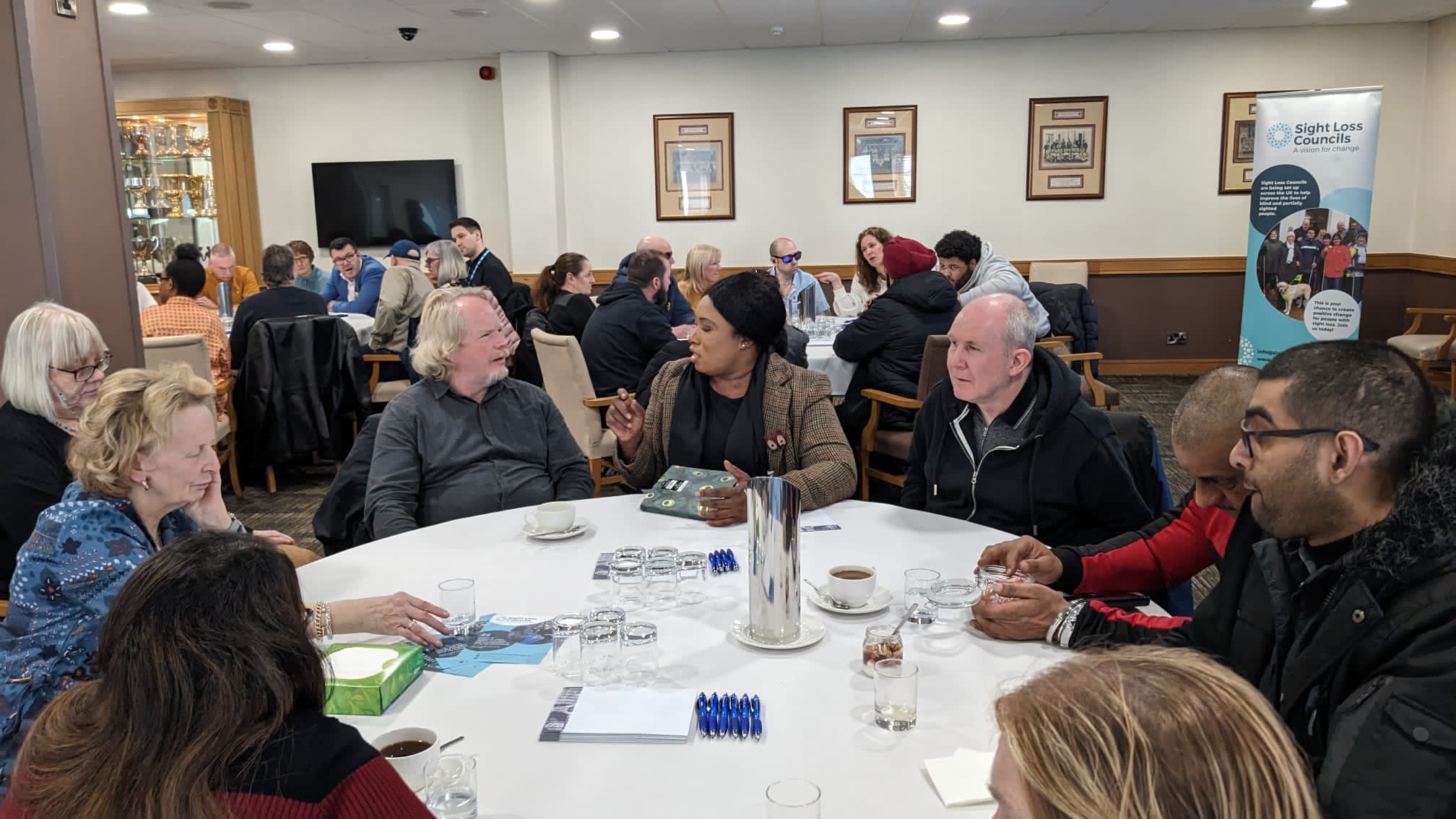 Table discussions with blind and partially sighted delegates, Sight Loss Council members and Beanca Mpofu (Vulnerabilities Team, University Hospitals Birmingham). Delegates are sat around circular tables in a large room. A Sight Loss Council banner is in the background.