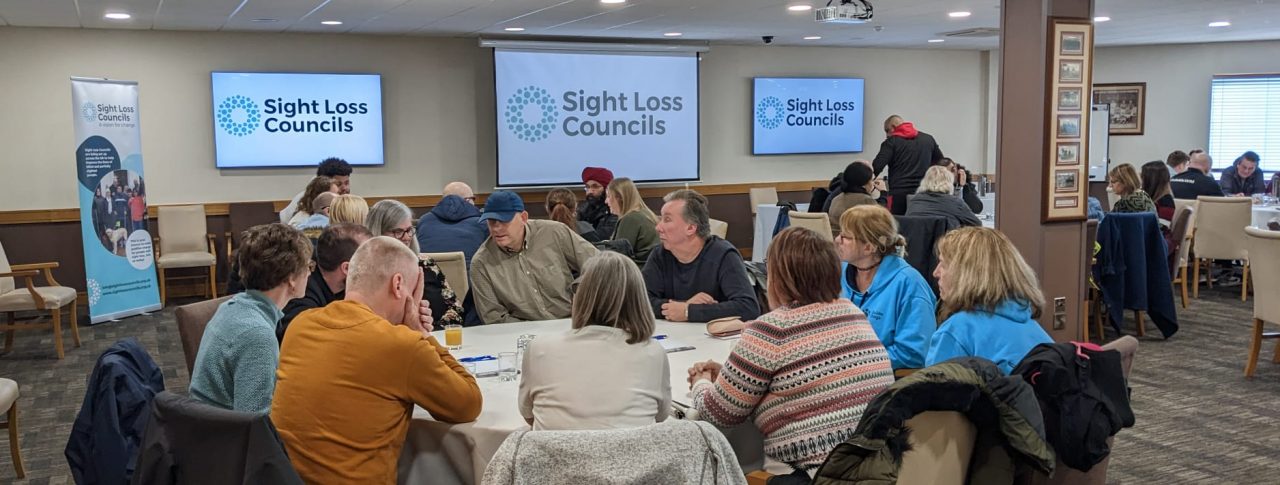 able discussions with blind and partially sighted delegates, Sight Loss Council members and Jamie Reilly (Vulnerabilities Team, University Hospitals Birmingham). Delegates are sat around circular tables in a large room. Screens and a banner displaying the Sight Loss Council logo are in the background.