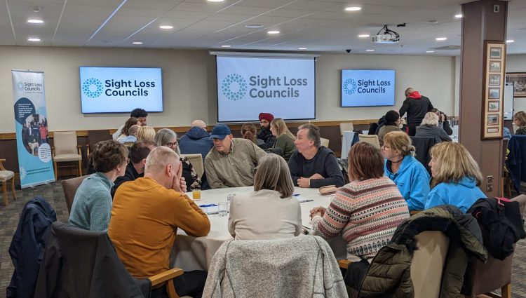 able discussions with blind and partially sighted delegates, Sight Loss Council members and Jamie Reilly (Vulnerabilities Team, University Hospitals Birmingham). Delegates are sat around circular tables in a large room. Screens and a banner displaying the Sight Loss Council logo are in the background.