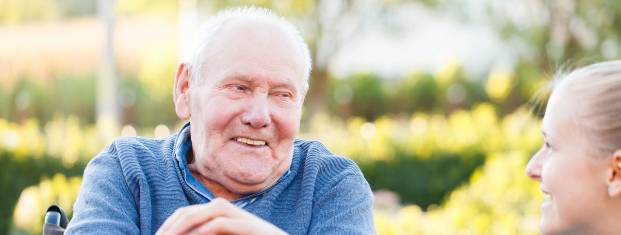 Close up of an elderly man sitting down. He has his hands on a walking stick. He is looking at a female carer, whose hand is on top of his, and they are smiling at each other.