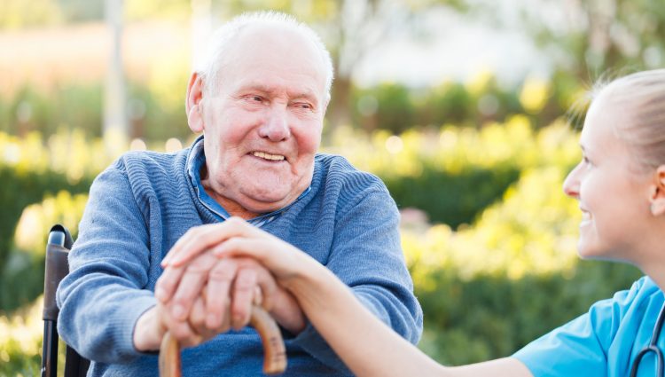 Close up of an elderly man sitting down. He has his hands on a walking stick. He is looking at a female carer, whose hand is on top of his, and they are smiling at each other.
