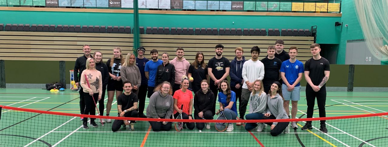 Merseyside SLC members and Martin Symcox, with students at Hope University Liverpool. They are standing on an indoor tennis court, behind the net. They are all looking at the camera and smiling.