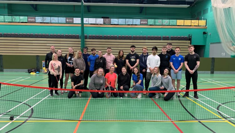 Merseyside SLC members and Martin Symcox, with students at Hope University Liverpool. They are standing on an indoor tennis court, behind the net. They are all looking at the camera and smiling.