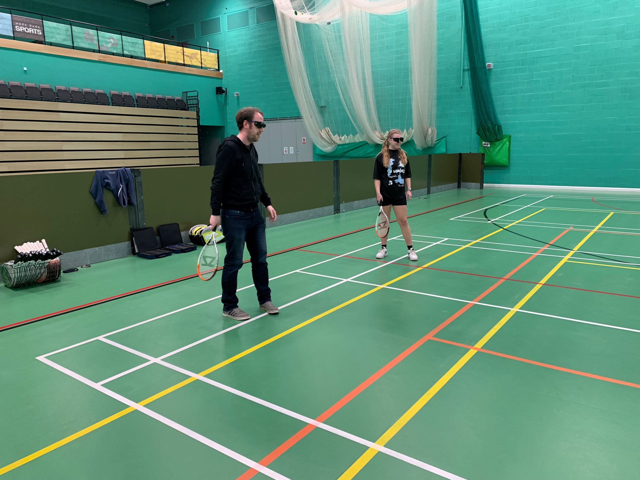 Image of two sports students on the indoor tennis court. Both are wearing sim specs and holding a tennis racket.