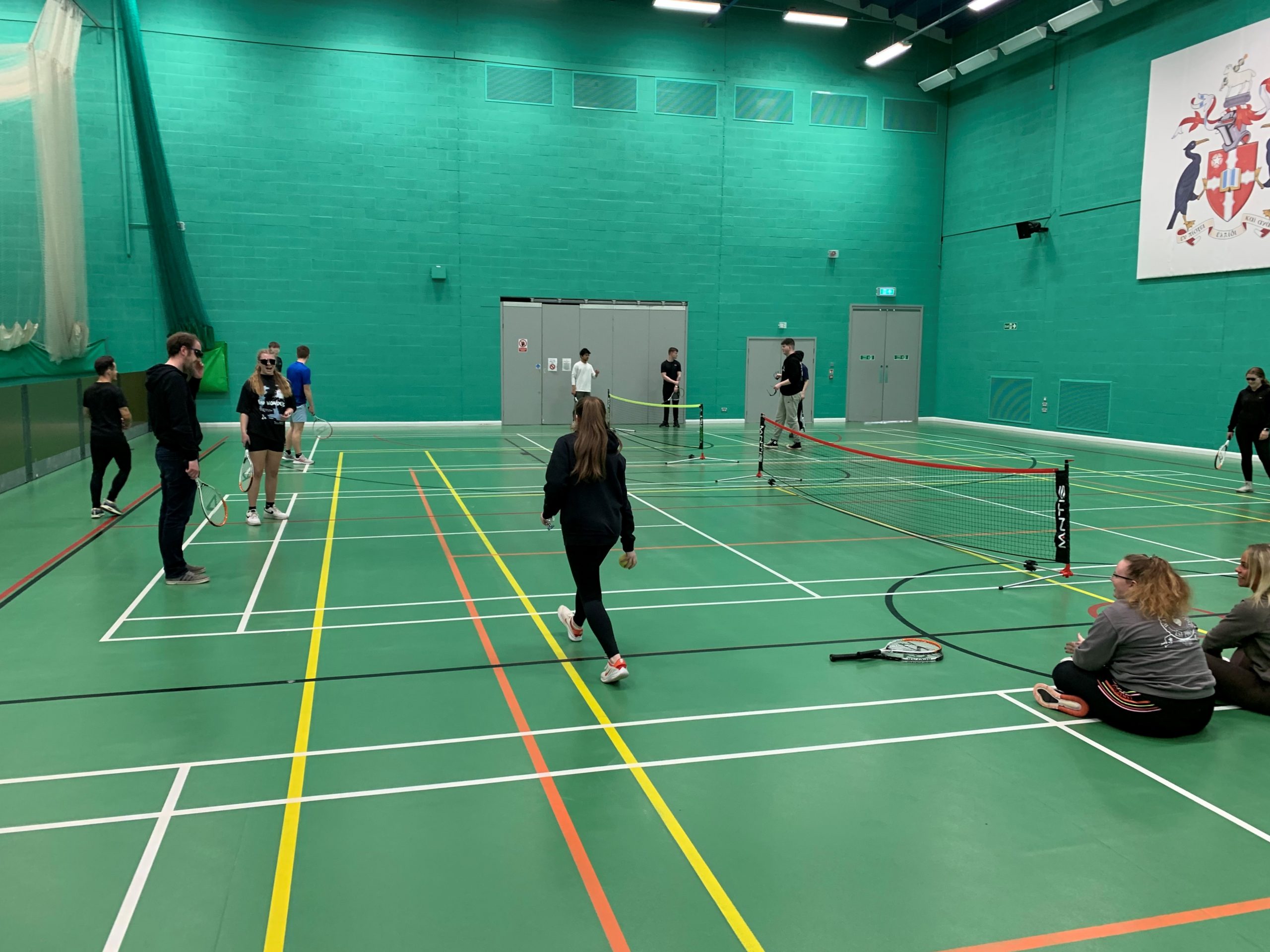 Image looks across the indoor tennis courts. Sports students are shown during a VI tennis session. Some are wearing sim specs whilst they play tennis.