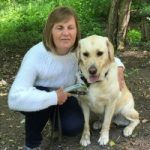 Julie Stephens, Gloucestershire SLC member, is knelt next to her guide dog, Heidi. She is on a woodland path with trees behind her.