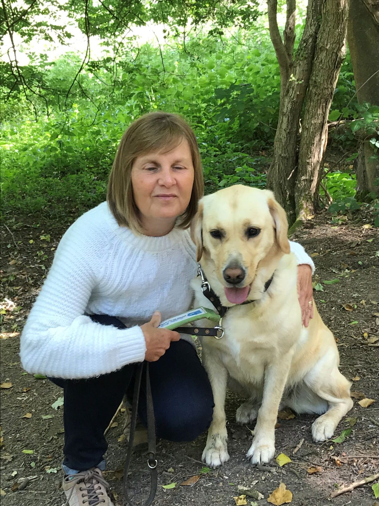 Julie Stephens, Gloucestershire SLC member, is knelt next to her guide dog, Heidi. She is on a woodland path with trees behind her.