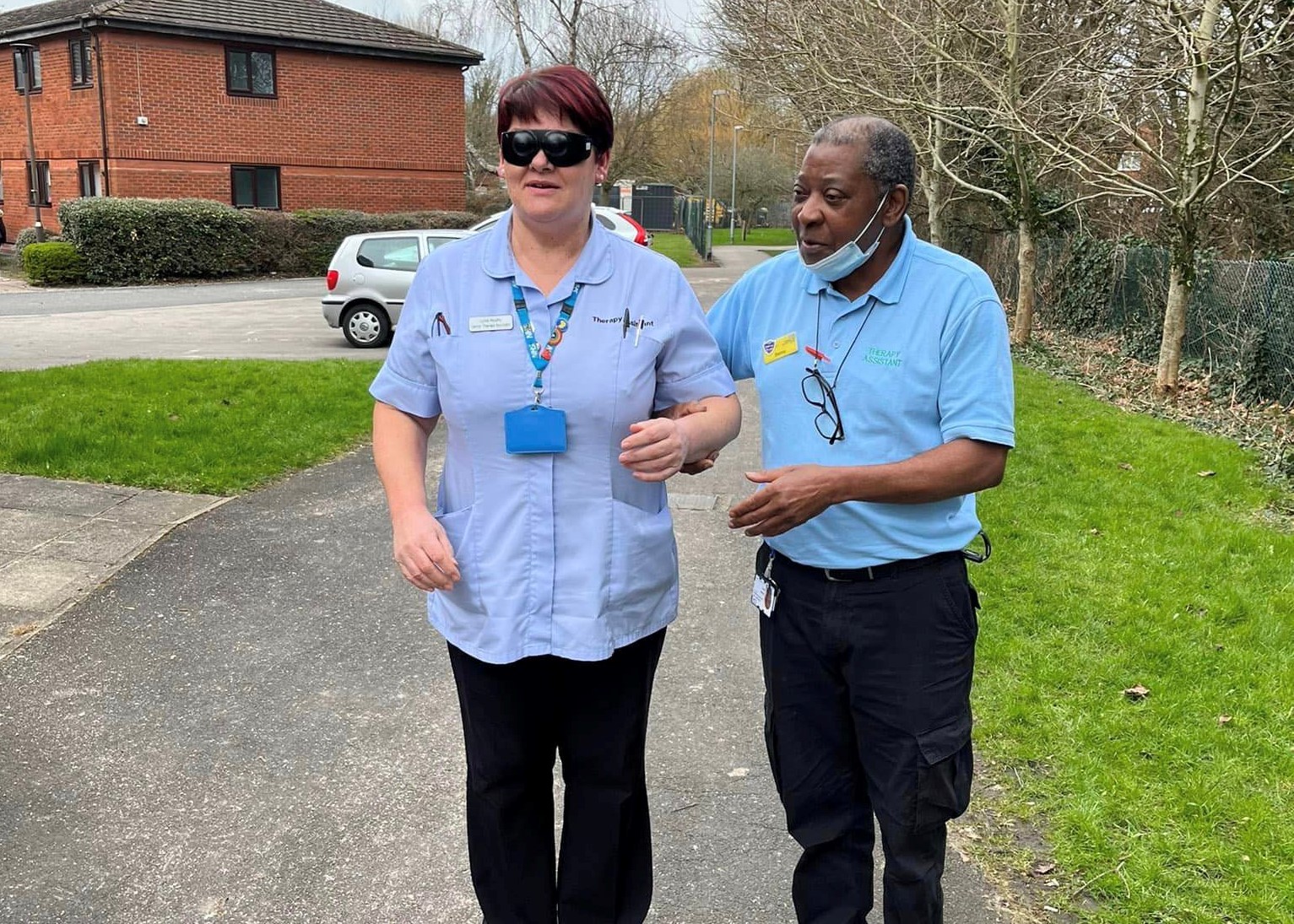 Image shows a male and female member of staff, taking part in the sighted guide training. The man is guiding the lady by the elbow. They are outside.