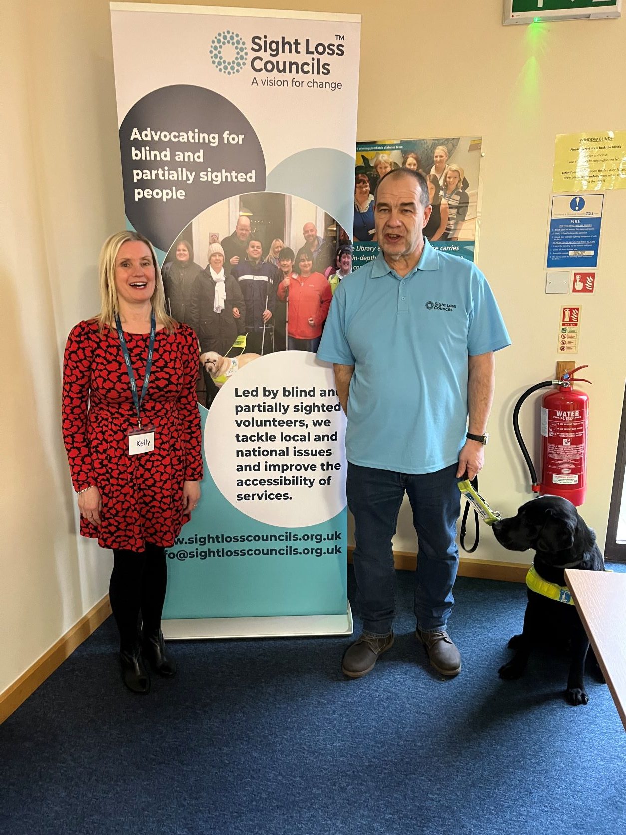 Kelly Barton, engagement manager for the north east, and Merseyside SLC member, Mick, standing in front of the Sight Loss Councils banner. Mick's guide dog Kip is sitting next to Mick.