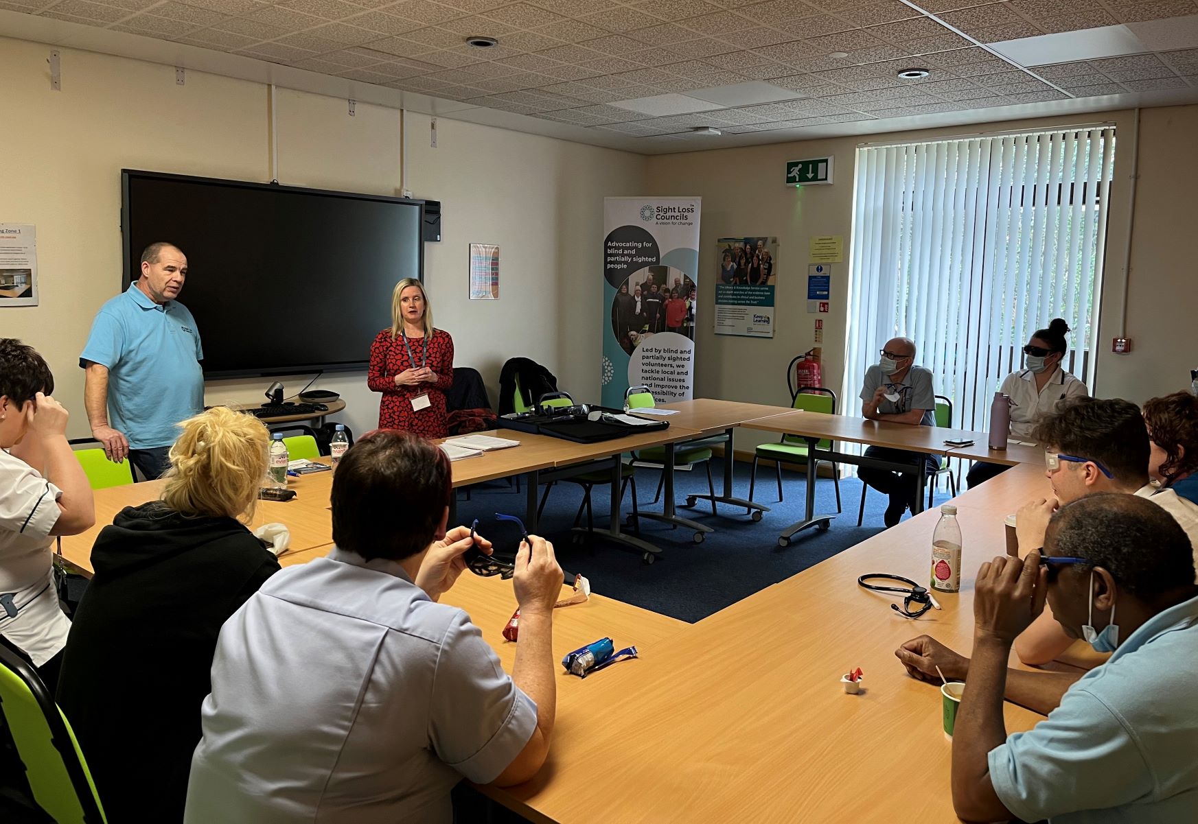 Kelly Barton and Mick Spriggs are presenting to staff members, who are seated and trying on simulation spectacles.