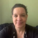 Denise, London SLC member, is seated, facing the camera, and smiling. Her hair is tied back.