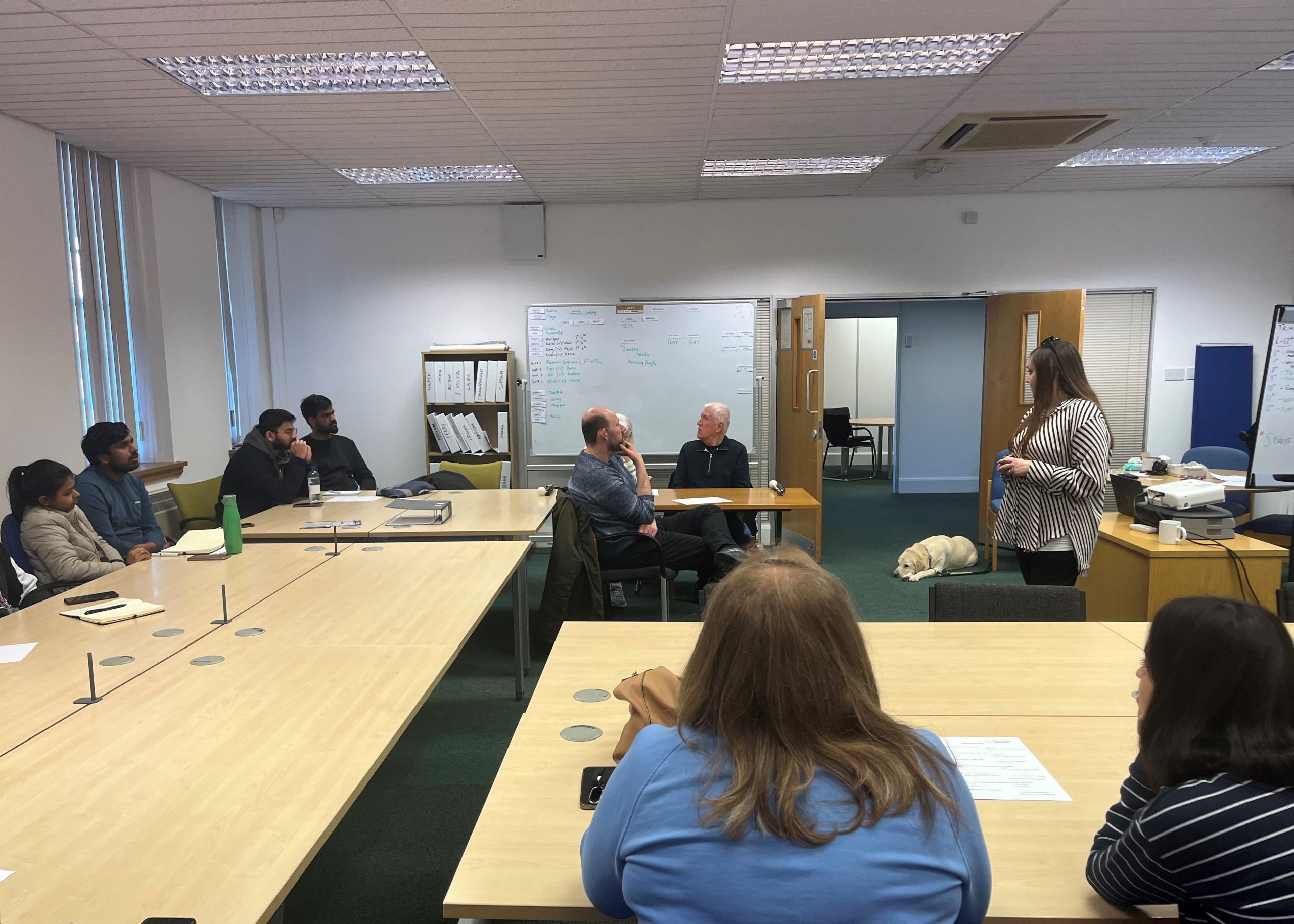 Engagement Manager, Samantha, is stood in front of Ethos Farm Staff, presenting the vision awareness session. Guide dog Lizzie is laying on the floor near her feet.