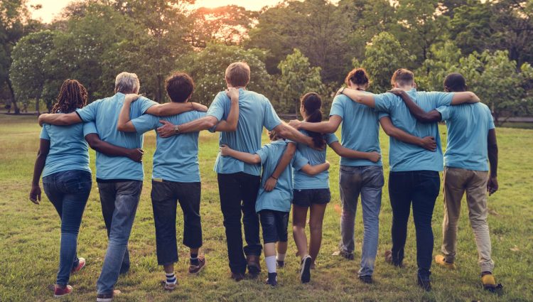 Group of volunteers with their arms around each other, facing away from the camera. They are outside and are all wearing light blue t-shirts.