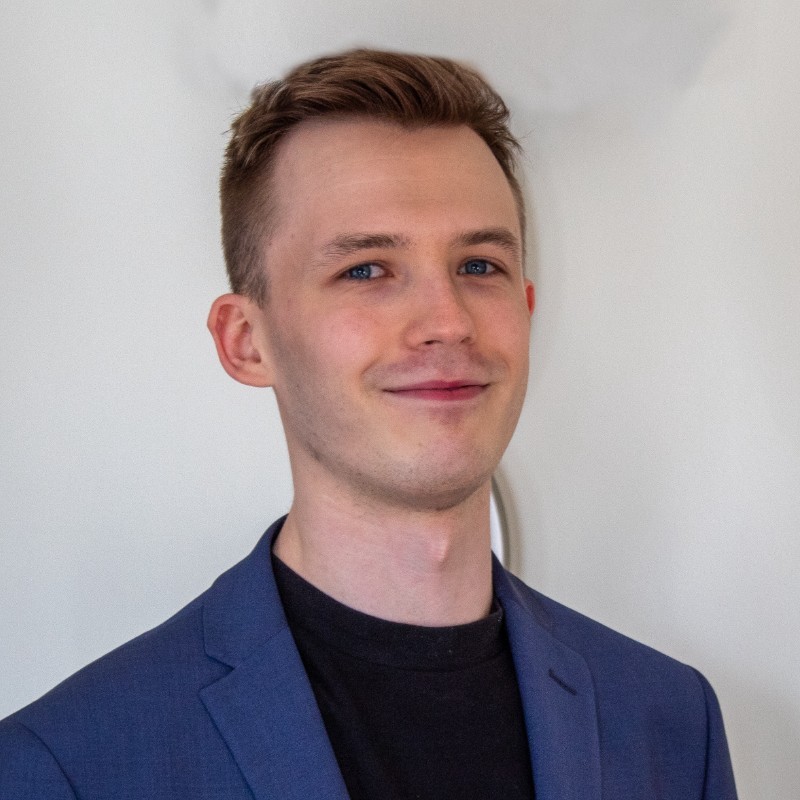 Headshot of Lewis Oldroyd, West Yorkshire SLC member. Lewis is standing against a white wall. He is wearing a blue suit jacket and black t-shirt. He is smiling at the camera.