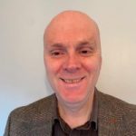 Head shot of Matt Harrison, Engagement Manager for East Midlands. Matt is standing against a white wall and smiling at the camera. He is wearing a black shirt and a blazer.