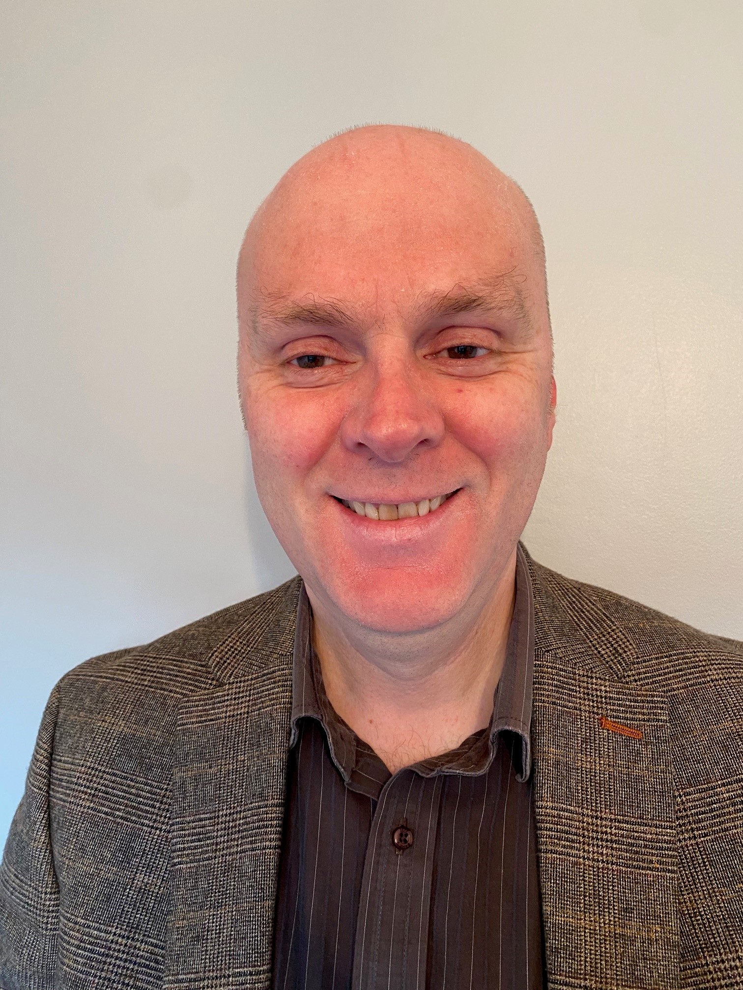 Head shot of Matt Harrison, Engagement Manager for East Midlands. Matt is standing against a white wall and smiling at the camera. He is wearing a black shirt and a blazer.