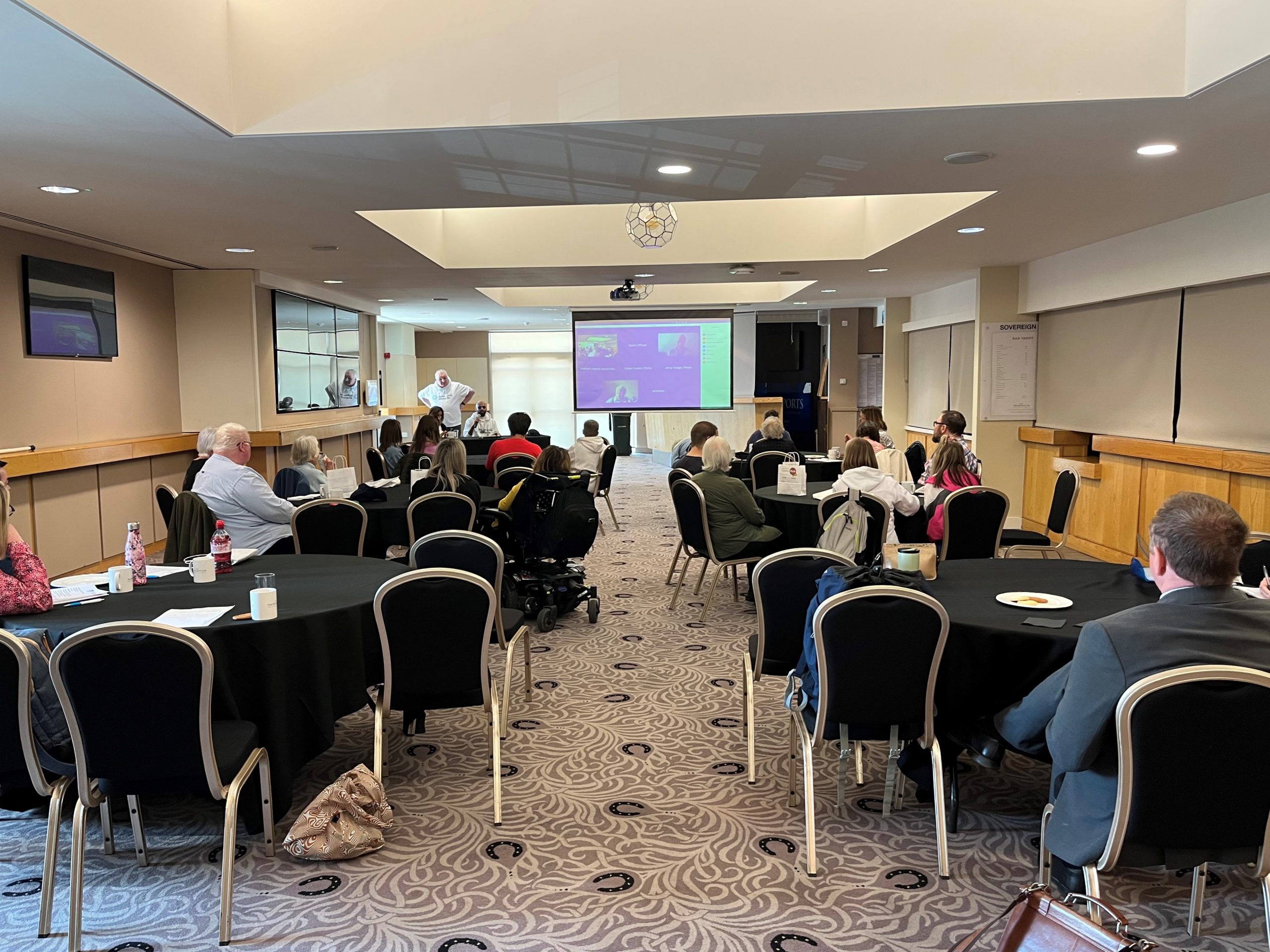 Delegates sitting around tables at Bristol & Gloucestershire SLC event. The image is taken from the back of the room, looking towards a large overhead projector. Alun Davies, Engagement Manager for the South West, is standing at the front of the room, introducing the event.