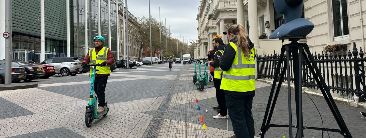 Someone on an e-scooter on a pedestrianised road. Three members of PEARL UCL are standing on the edge with their equipment, monitoring the experiment.