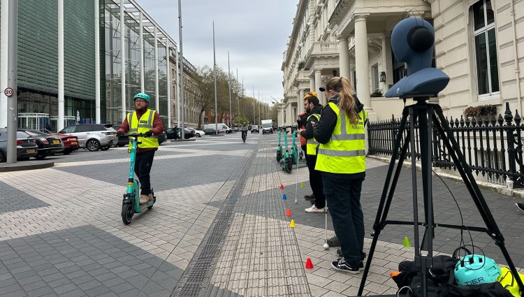 Someone on an e-scooter on a pedestrianised road. Three members of PEARL UCL are standing on the edge with their equipment, monitoring the experiment.