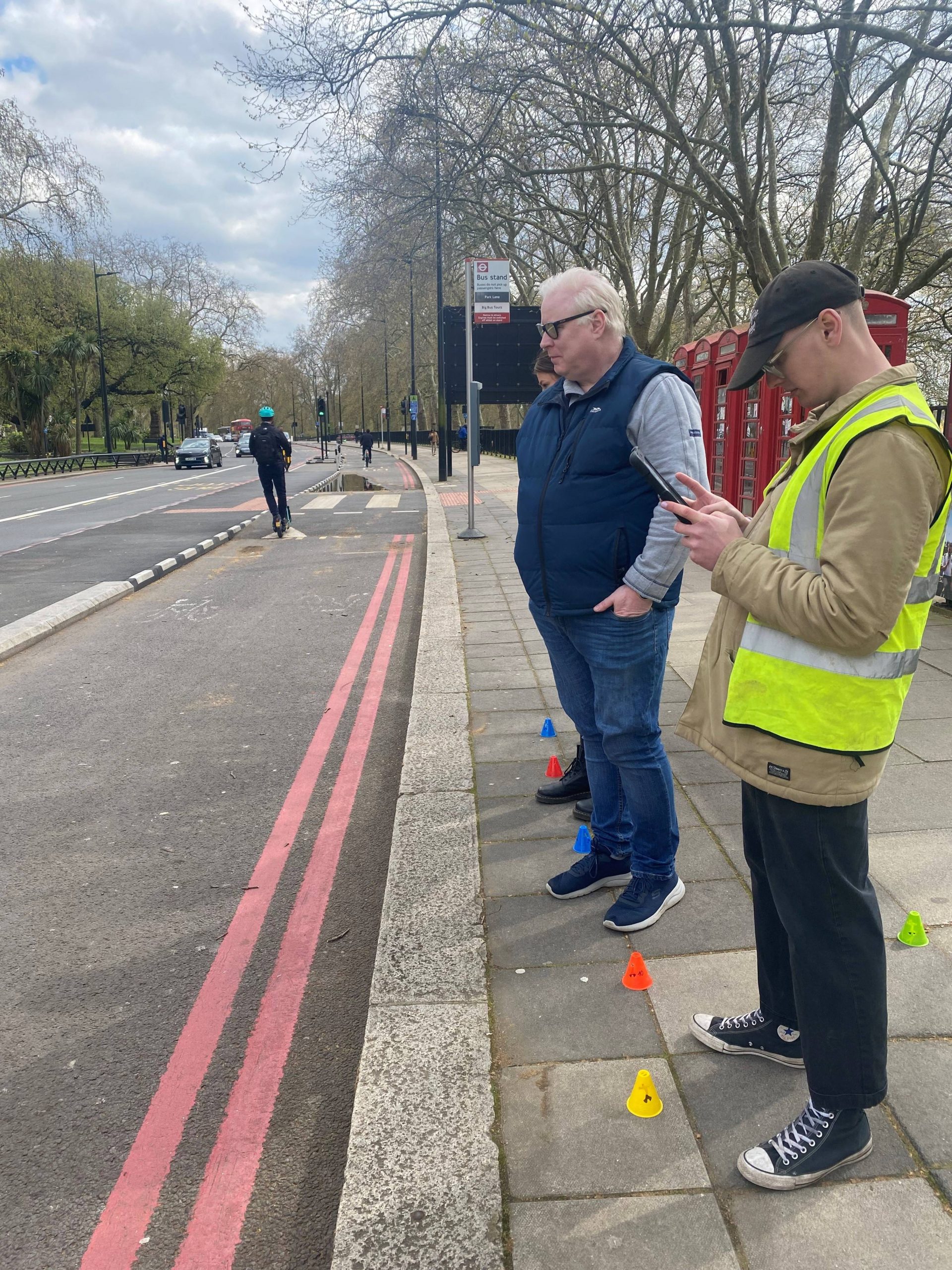 Senior Engagement Manager, Iain Mitchell, standing on a path next to a road with a member of the AVA testing team from UCL Pearl. Iain is listening out for an approaching e-scooter.