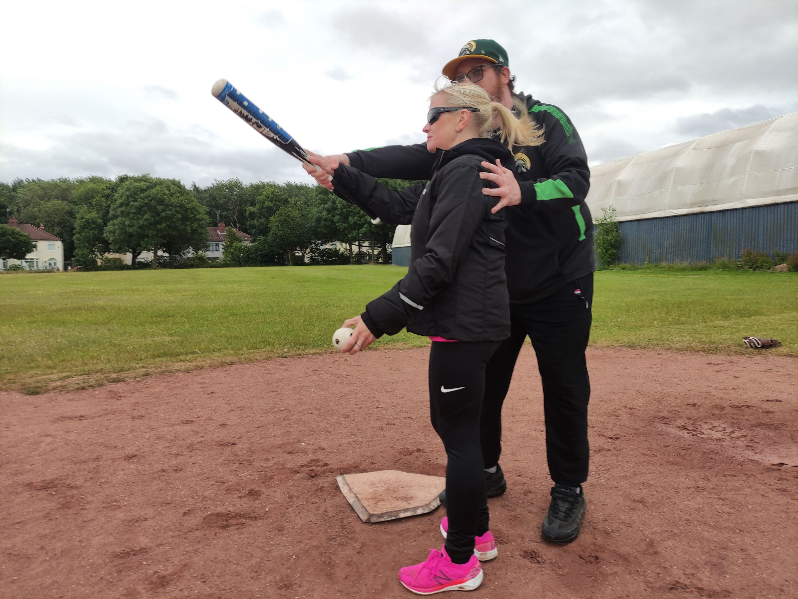 Kelly Barton, Engagement Manager for the North East during a VI Baseball session. She is standing on the field at the home base. She is with an instructor who is guiding her where to position the bat. Kelly is holding the baseball in her other hand.