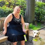 Leanne, London SLC member is sitting on a park bench with her guide dog. She is looking forwards, laughing.