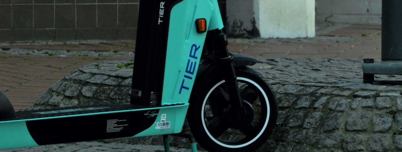 A close up of the front wheel of a TIER e-scooter. The e-scooter is turquoise, with TIER written on the frame, by the wheel.