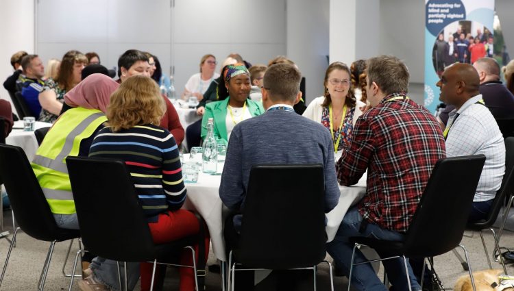 Blind and partially sighted volunteers are sat around circular tables having a discussion at a volunteer event. A Sight Loss Council banner is in the background.