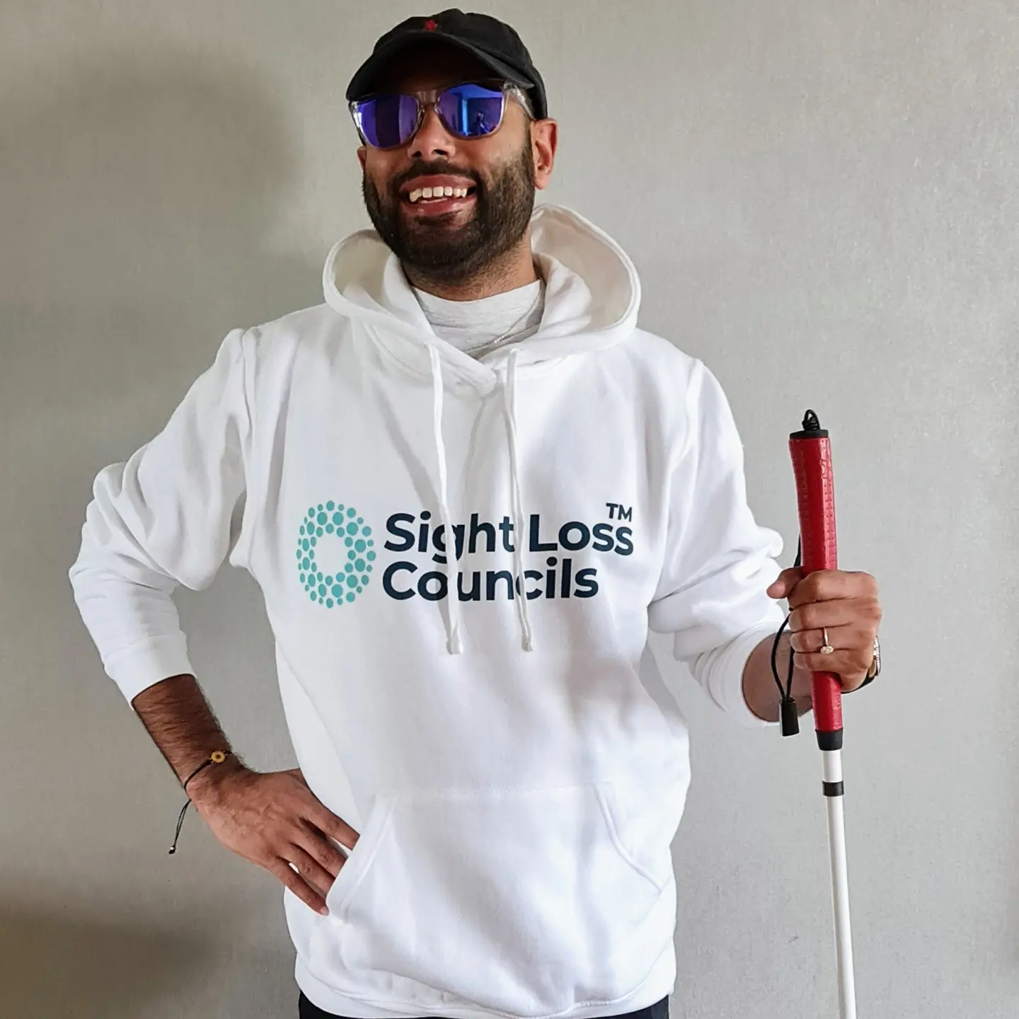 Yahya Pandor, Gloucestershire SLC member, is standing against a white wall, smiling at the camera. Yahya is wearing a baseball cap and sunglasses. He is wearing a white 'Sight Loss Councils' hoodie. he has one hand on his hip and the other is holding his cane up.