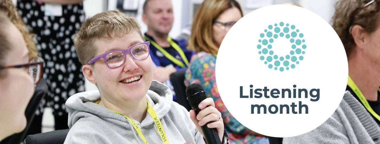 A Sight Loss Council Volunteer, Emma, is sat at a volunteer event. She is holding the microphone getting ready to speak. She is smiling. Text says 'Listening Month' and has the Sight Loss Council logo icon above it.