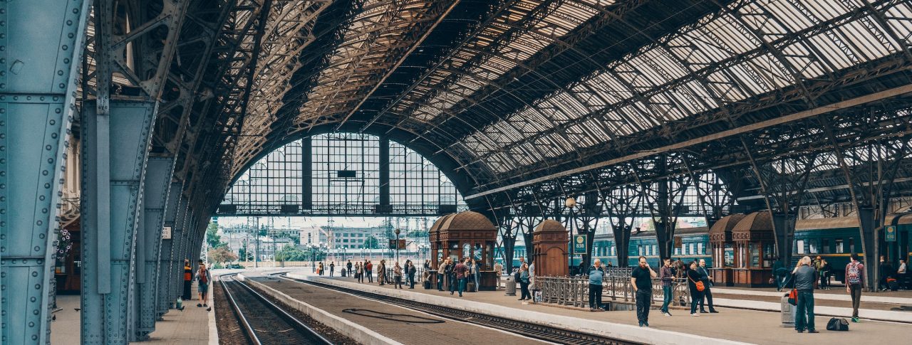 A Victorian rail station with a domed glass roof and ornate platform pillars. Several passengers are on the platform in the distance, waiting for a train.