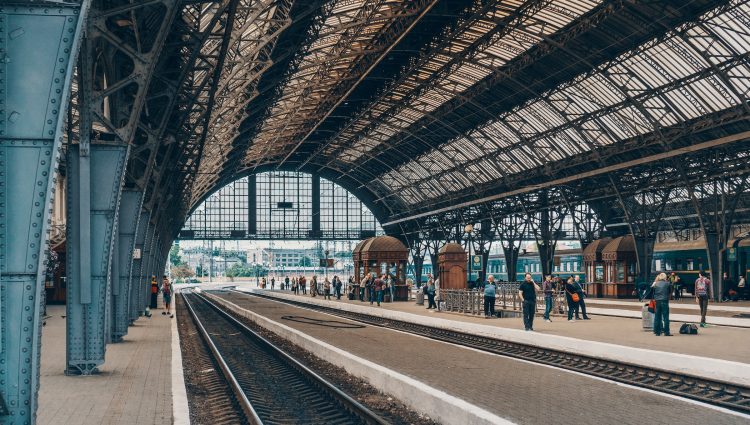 A Victorian rail station with a domed glass roof and ornate platform pillars. Several passengers are on the platform in the distance, waiting for a train.