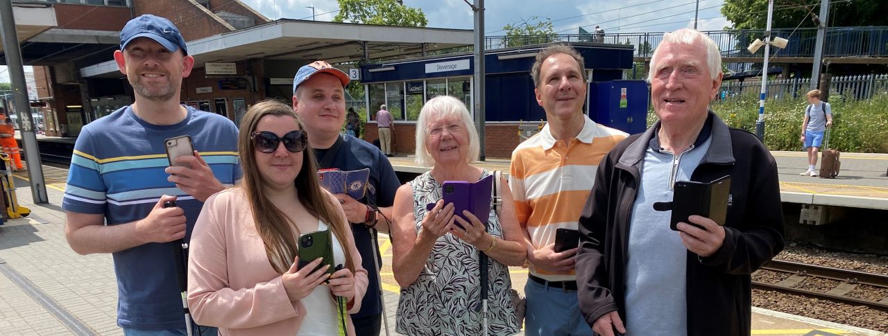 Members of Bedfordshire SLC are stood with Samantha Leftwich, Engagement Manager for East England. They are stood on a platform at Stevenage train station as part of the Aira trial. They are all holding their smartphones in one hand, their long cane in the other.