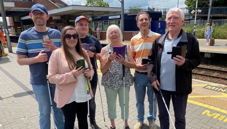 Members of Bedfordshire SLC are stood with Samantha Leftwich, Engagement Manager for East England. They are stood on a platform at Stevenage train station as part of the Aira trial. They are all holding their smartphones in one hand, their long cane in the other.