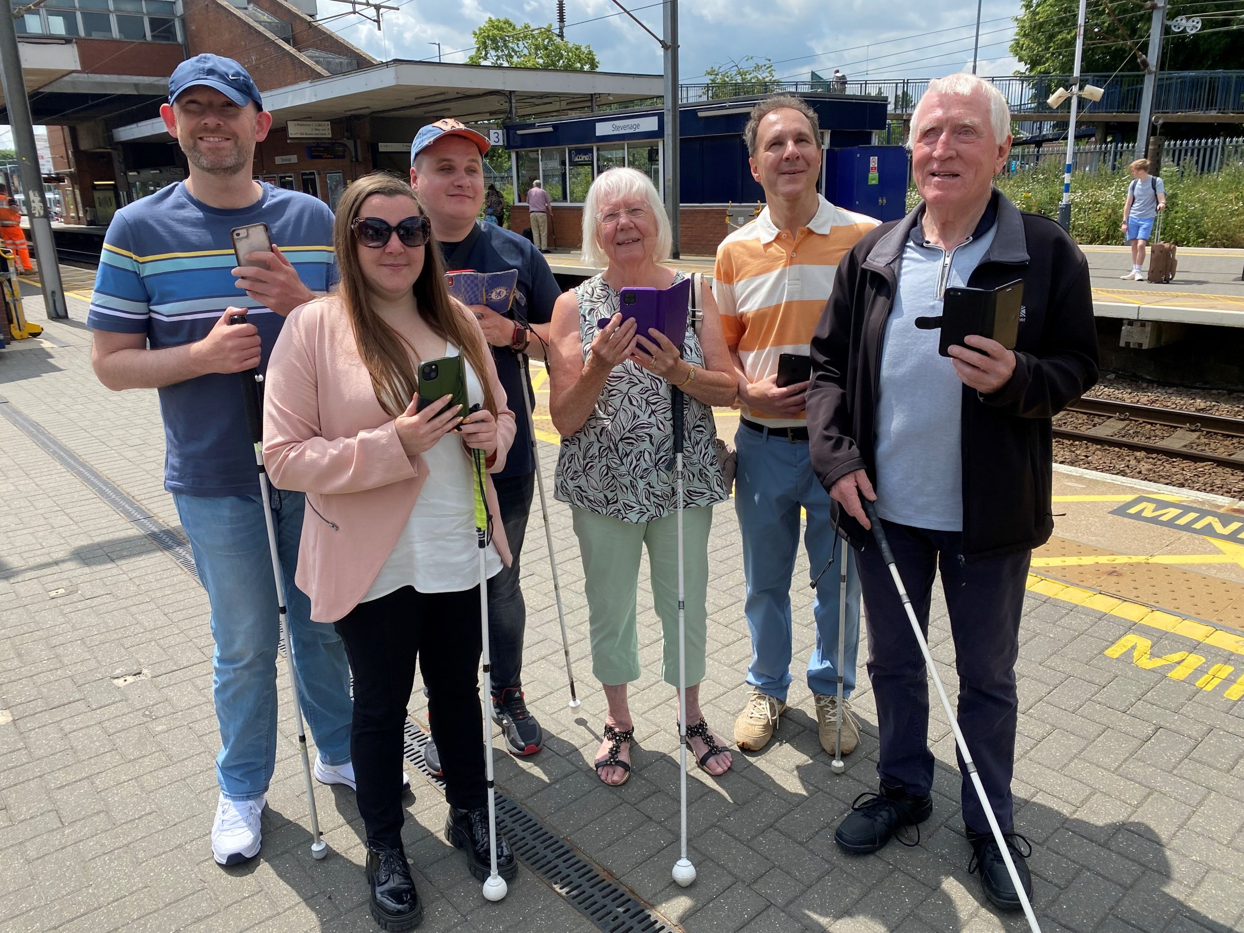 Members of Bedfordshire SLC are stood with Samantha Leftwich, Engagement Manager for East England. They are stood on a platform at Stevenage train station as part of the Aira trial. They are all holding their smartphones in one hand, their long cane in the other.