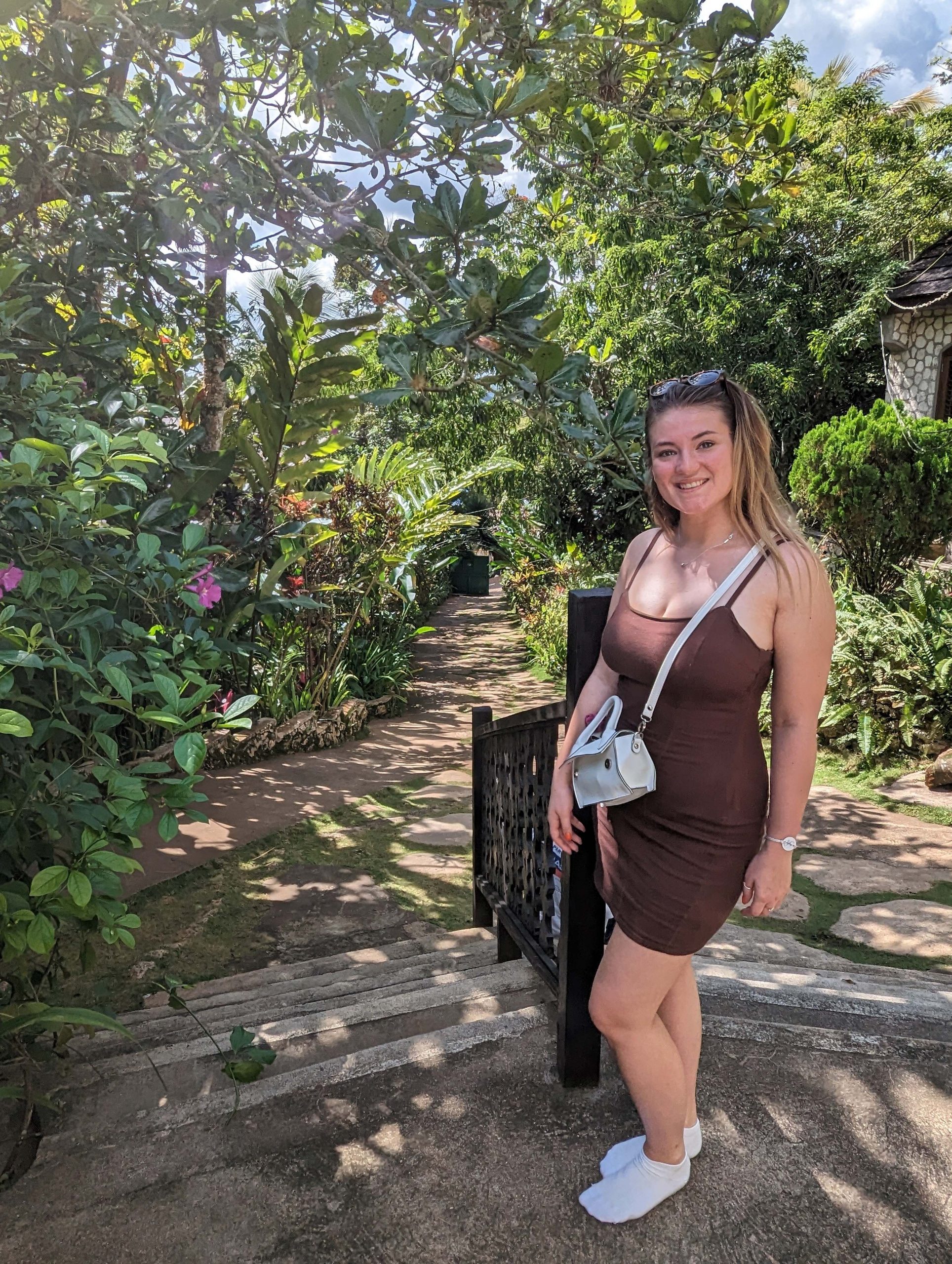 Belle during a holiday to Jamaica in 2022. She is wearing a brown sundress and is standing at the entrance of a pathway, surrounded by green trees and shrubs.