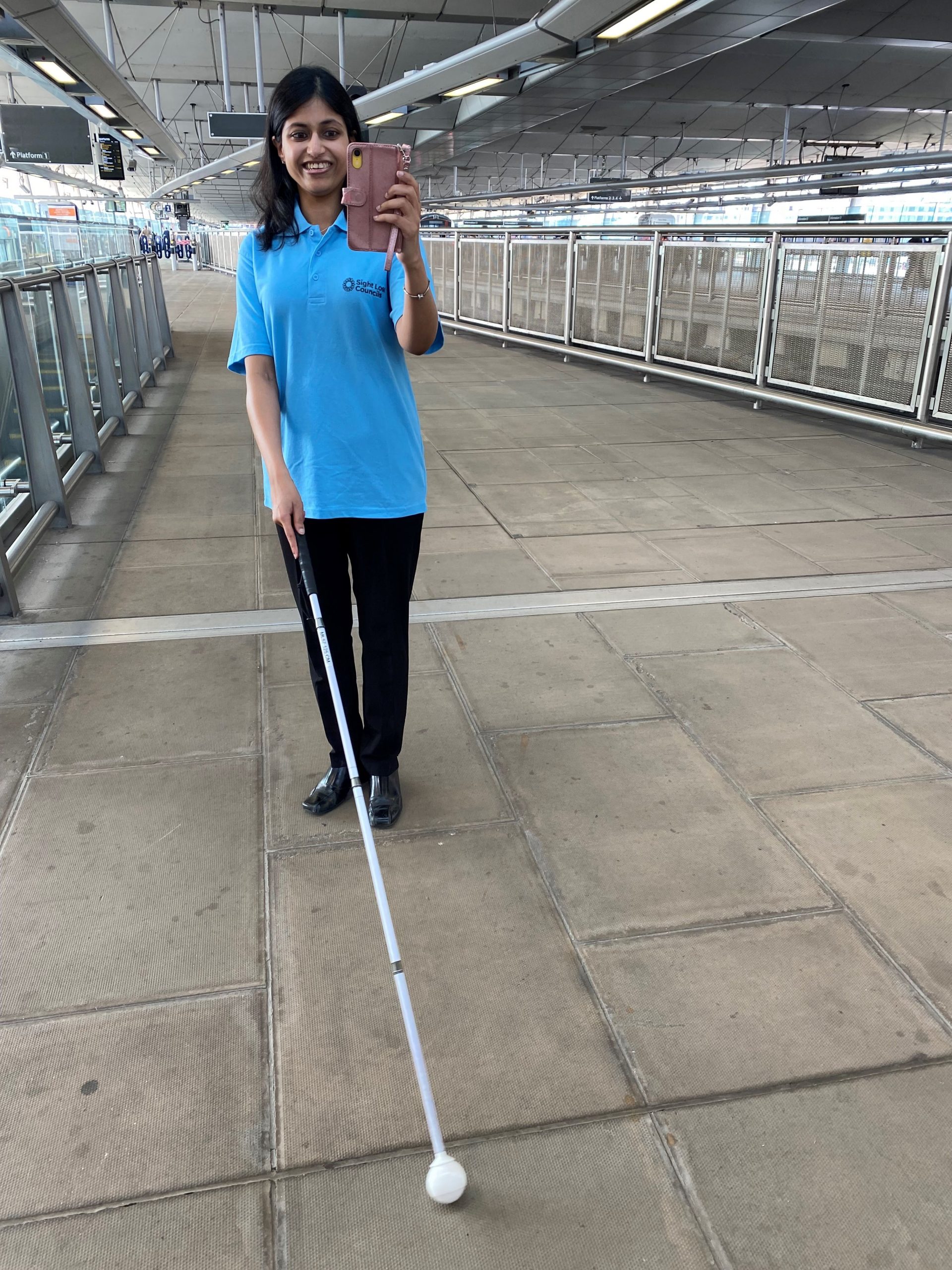 Vidya, London SLC member, is walking along a platform at London Blackfriars. She is holding her phone up in one hand and using her long cane in the other. She is smiling.