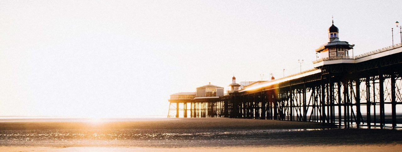 Blackpool pier, taken from the sandy beach below it. The image is taken at sunrise, and sunlight floods the image. It is low tide, so the cast iron structure of the pier is visible, with a couple of ornate buildings shown on top.