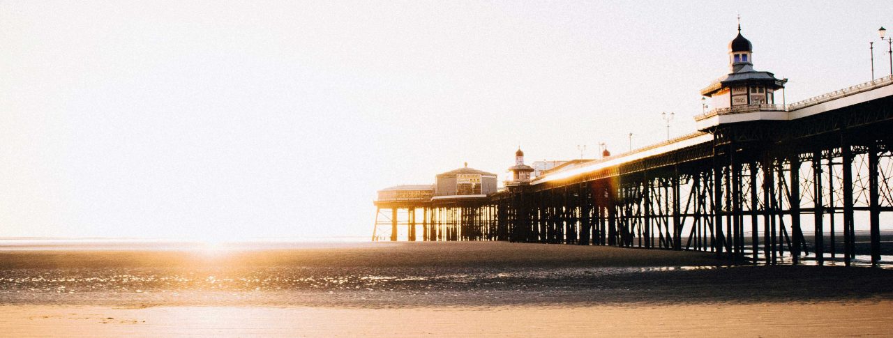 Blackpool pier, taken from the sandy beach below it. The image is taken at sunrise, and sunlight floods the image. It is low tide, so the cast iron structure of the pier is visible, with a couple of ornate buildings shown on top.