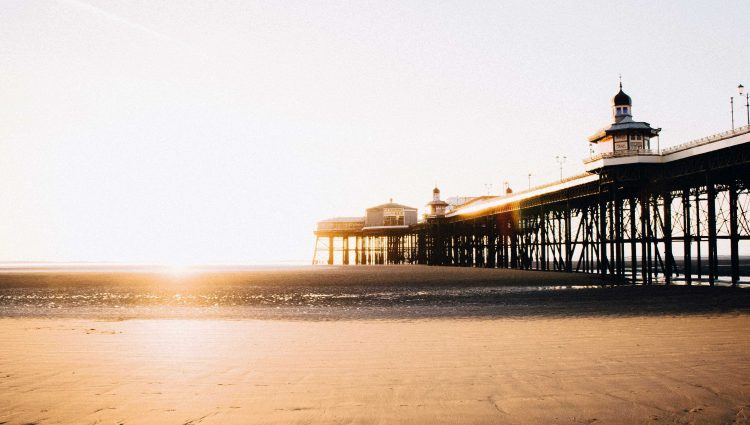 Blackpool pier, taken from the sandy beach below it. The image is taken at sunrise, and sunlight floods the image. It is low tide, so the cast iron structure of the pier is visible, with a couple of ornate buildings shown on top.